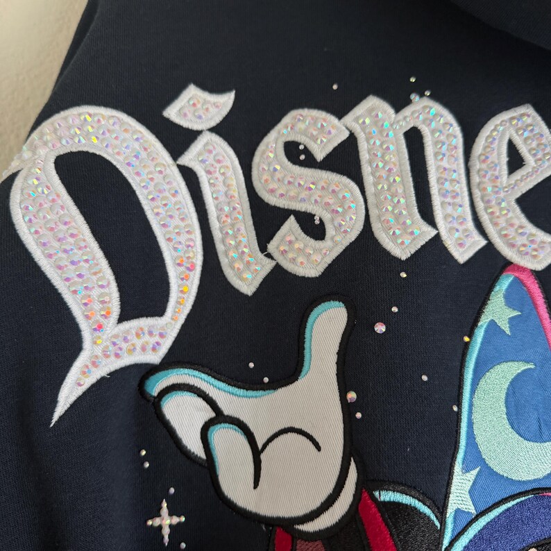 May include: A dark navy blue hoodie featuring the word Disney embroidered in white with iridescent rhinestone embellishments. Below the text is a white gloved hand and a blue wizard hat with a crescent moon and star pattern, accented by rhinestones.