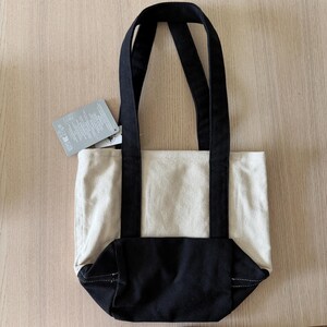 Op de afbeelding: Een canvas draagtas met twee tinten. Het bovenste gedeelte is naturel beige, terwijl de bodem en handvatten zwart zijn. Er is een label bevestigd. De tas is ontworpen om items te dragen. Afmetingen niet zichtbaar.