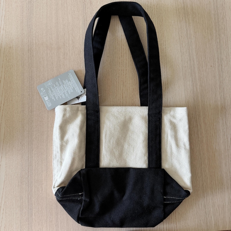 Op de afbeelding: Een canvas draagtas met twee tinten. Het bovenste gedeelte is naturel beige, terwijl de bodem en handvatten zwart zijn. Er is een label bevestigd. De tas is ontworpen om items te dragen. Afmetingen niet zichtbaar.