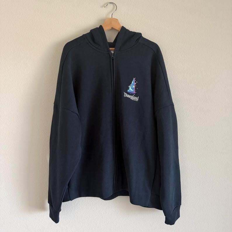 May include: A black zip-up hoodie featuring a hood, long sleeves, and a front zipper closure. On the upper left chest, there is a graphic of a blue wizard hat with a crescent moon and stars, accompanied by the text Disneyland Resort in white.