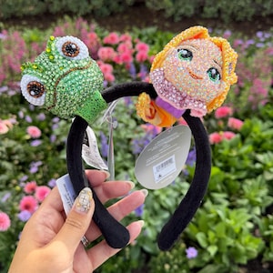 May include: A black headband featuring two plush character figures covered in iridescent rhinestones. On the left is a green chameleon with large eyes, and on the right is a blonde character with a pink collar. Both figures have attached tags.