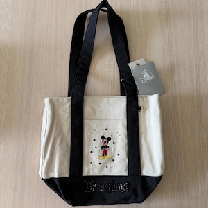 Op de afbeelding: Canvas tote bag met zwarte handvatten en basis. Voorzien van een Mickey Mouse-afbeelding met een spreiding van stippen en het woord "Disneyland" in een kralenontwerp. Inclusief een Disney-tag.