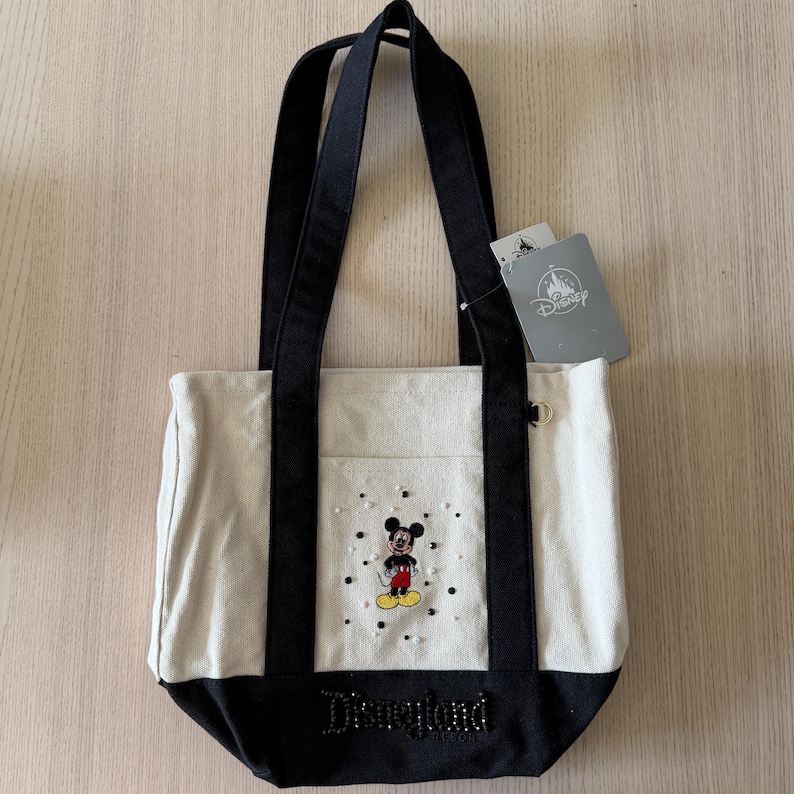 Op de afbeelding: Canvas tote bag met zwarte handvatten en basis. Voorzien van een Mickey Mouse-afbeelding met een spreiding van stippen en het woord "Disneyland" in een kralenontwerp. Inclusief een Disney-tag.