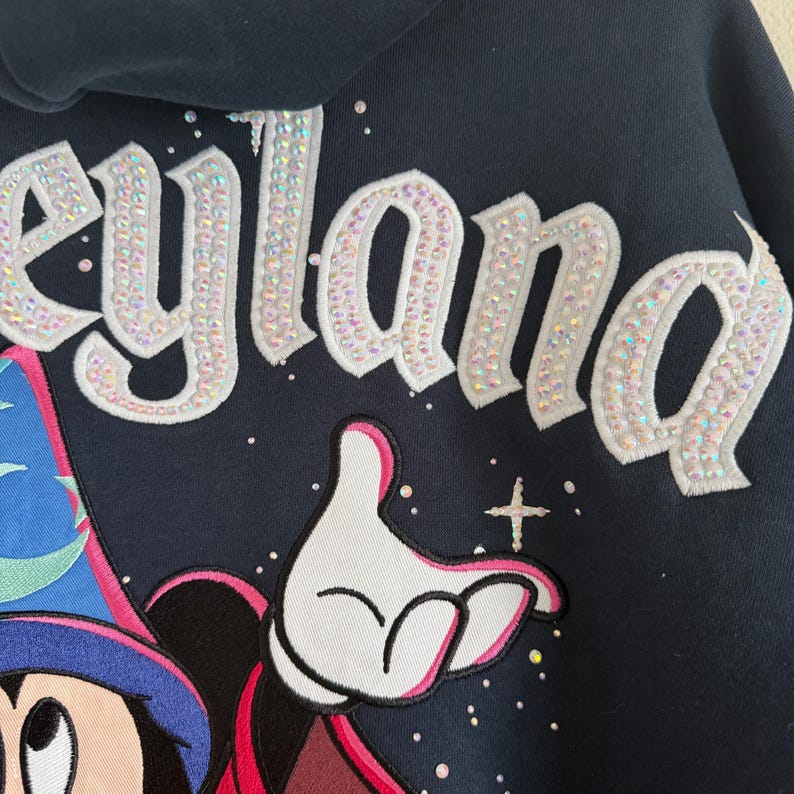May include: A dark navy blue hooded sweatshirt featuring an embroidered graphic of a character wearing a blue wizard hat. The text Disneyland is displayed in large, white, fabric-stitched letters embellished with iridescent, sparkling rhinestones.