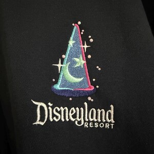May include: A black fabric garment featuring an embroidered blue wizard hat with a light green crescent moon and star design. The embroidery includes small white star accents and scattered beads. The text Disneyland RESORT is stitched in white.