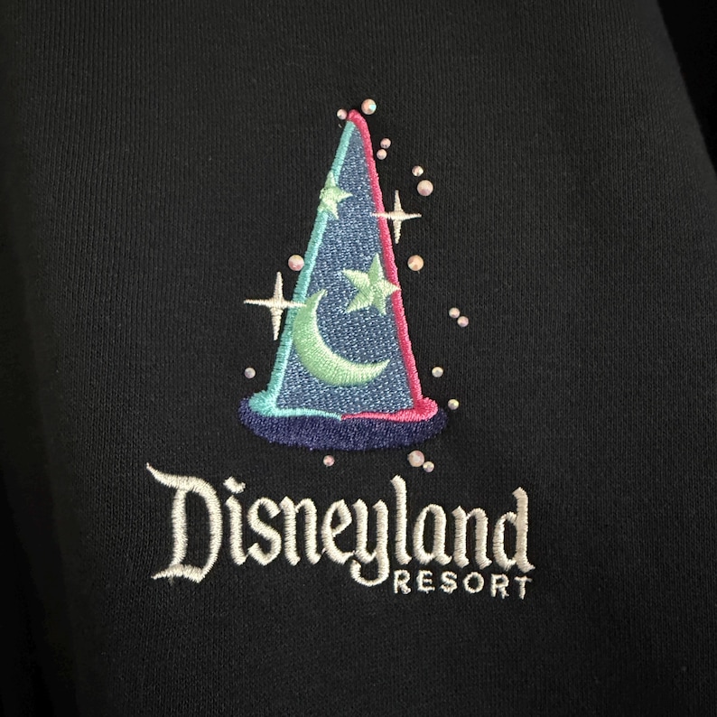 May include: A black fabric garment featuring an embroidered blue wizard hat with a light green crescent moon and star design. The embroidery includes small white star accents and scattered beads. The text Disneyland RESORT is stitched in white.
