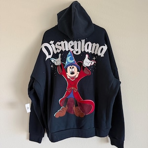 May include: A black hooded sweatshirt featuring a large graphic of Mickey Mouse as a sorcerer on the back. The word Disneyland is printed in white above the character, who wears a blue hat with a moon and star, and a long red robe.