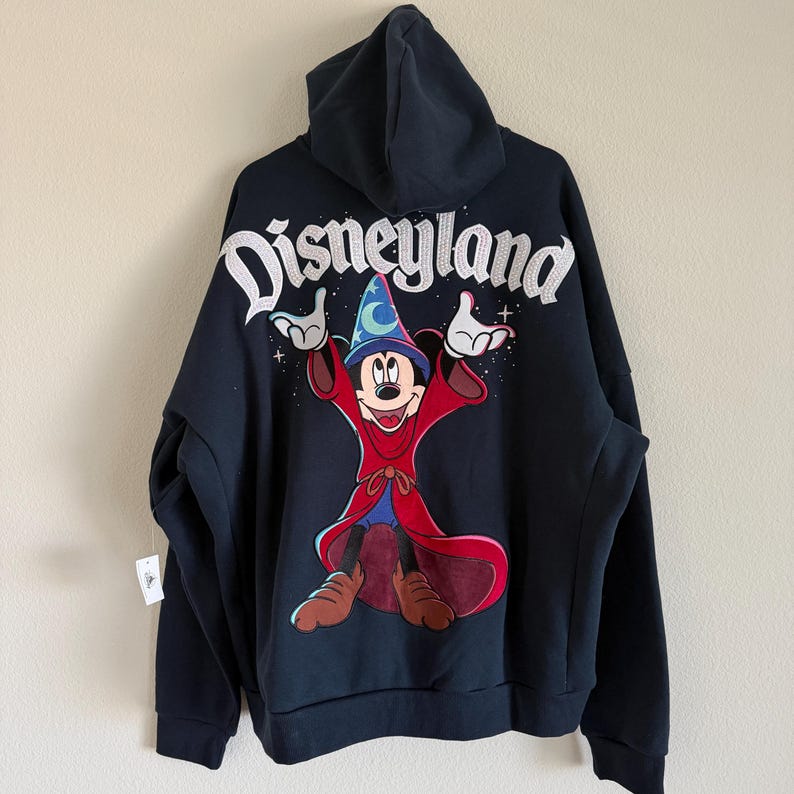 May include: A black hooded sweatshirt featuring a large graphic of Mickey Mouse as a sorcerer on the back. The word Disneyland is printed in white above the character, who wears a blue hat with a moon and star, and a long red robe.