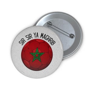 May include: A round pin-back button featuring a red soccer ball graphic with a green star in the center. The text "SIR SIR YA MAGHRIB" is printed in black in an arc above the ball. The button has a metallic back with a pin mechanism.