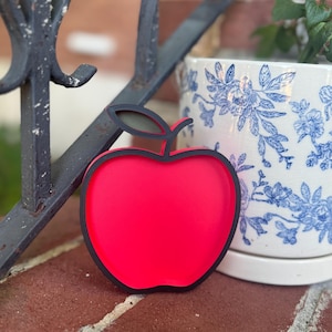May include: A bright red apple-shaped tray with a thick black border and a small black stem detail at the top. The tray has a smooth, flat surface and a raised edge, designed in the shape of a classic apple silhouette with a vibrant red center.