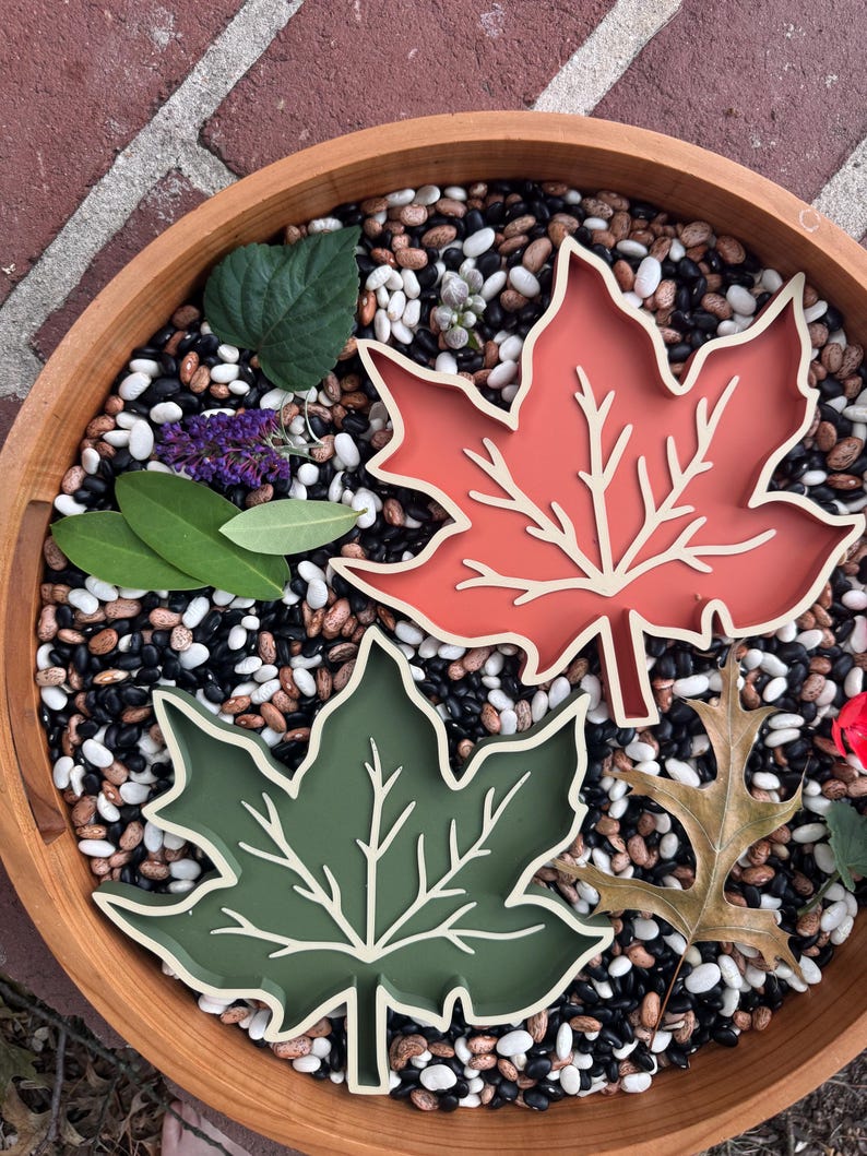 May include: Two leaf-shaped trays, one in a muted terracotta orange and the other in a deep olive green, each featuring a raised cream-colored vein pattern. The trays are arranged among scattered dried beans and several natural green leaves.