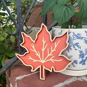 May include: A maple leaf-shaped decorative object featuring a solid terracotta-colored center with a raised, cream-colored outline and vein detailing. The item has a layered, dimensional construction with a smooth finish and a distinct stem at the base.