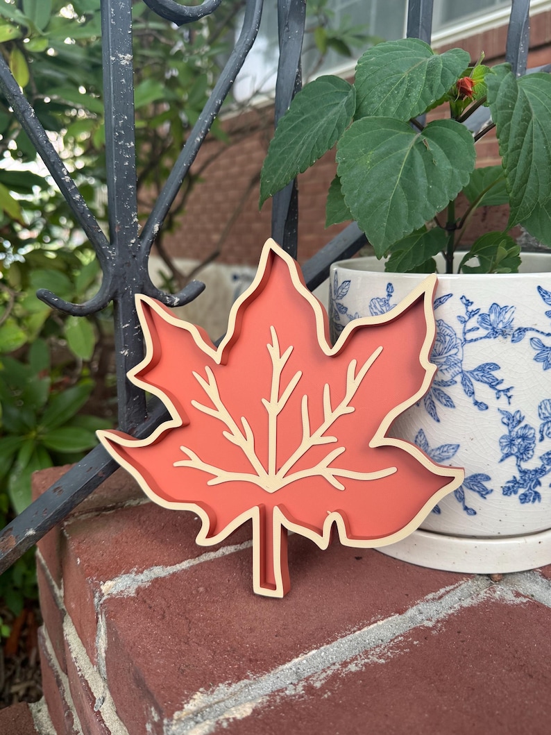 May include: A maple leaf-shaped decorative object featuring a solid terracotta-colored center with a raised, cream-colored outline and vein detailing. The item has a layered, dimensional construction with a smooth finish and a distinct stem at the base.