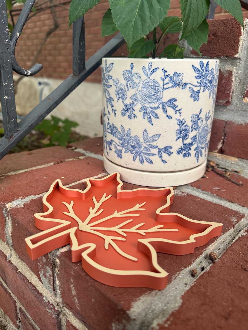 May include: A maple leaf-shaped tray in a solid burnt orange color with raised edges and a recessed vein pattern in a lighter cream tone. The tray is positioned in front of a cylindrical ceramic planter featuring a blue and white floral pattern.