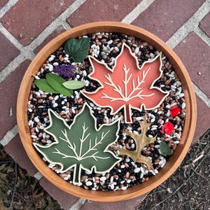 May include: A round wooden tray filled with a mixture of black, white, and brown dried beans. Inside the tray are two leaf-shaped decorative items, one in burnt orange and one in olive green, alongside a few small green leaves and red petals.