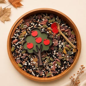 May include: A round wooden tray filled with black and white beans, scattered with colorful pasta shapes, a wooden spoon, and a dark green tree-shaped cutout featuring four bright red apple shapes attached to the branches.
