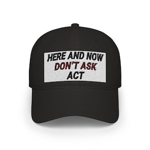 Peut inclure: Casquette de baseball noire avec un patch rectangulaire blanc. Le patch affiche le texte "HERE AND NOW DON'T ASK ACT" en noir et rouge. La casquette a une visière incurvée et une couronne structurée.