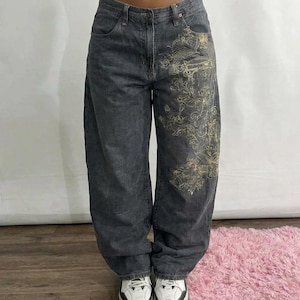 Peut inclure: Jean en denim gris foncé, coupe ample et ample. Le jean présente un motif floral brodé de couleur or clair sur le devant de la jambe gauche. Le modèle porte des baskets blanches et noires.