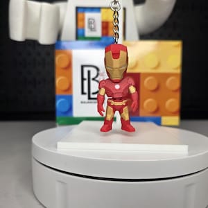 May include: A red and gold Iron Man keychain figurine. The miniature superhero figure is standing on a white platform. The background includes a colorful block design and a logo.