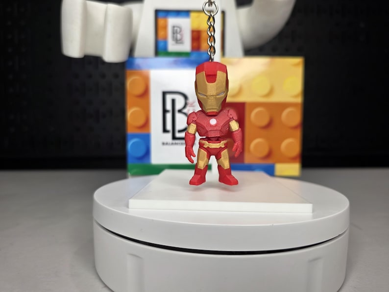 May include: A red and gold Iron Man keychain figurine. The miniature superhero figure is standing on a white platform. The background includes a colorful block design and a logo.