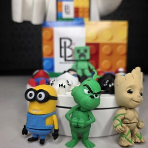 May include: A collection of colorful character figurines, including a yellow Minion, a green Grinch, and a tan Groot. Other figures include a green Minecraft Creeper and a red and blue Spider-Man. The figurines are arranged on a white platform.