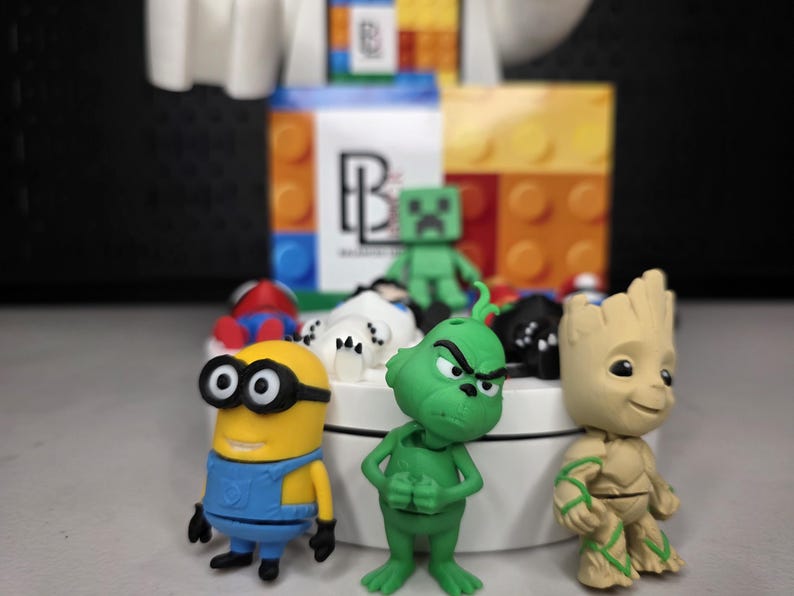 May include: A collection of colorful character figurines, including a yellow Minion, a green Grinch, and a tan Groot. Other figures include a green Minecraft Creeper and a red and blue Spider-Man. The figurines are arranged on a white platform.
