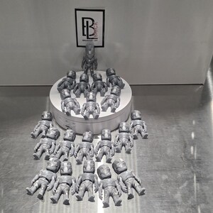 May include: A collection of twenty-one small, silver-colored, metallic-finish figurines arranged on and around a white circular display stand. Each figurine features a detailed, armored humanoid design. A logo with the letters BL is visible above.