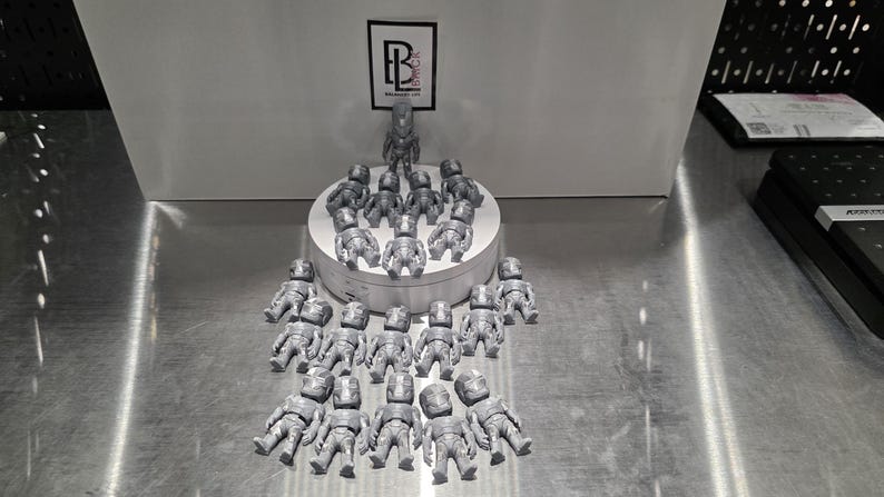 May include: A collection of twenty-one small, silver-colored, metallic-finish figurines arranged on and around a white circular display stand. Each figurine features a detailed, armored humanoid design. A logo with the letters BL is visible above.