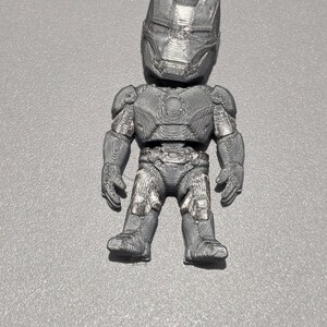 May include: A small, three-dimensional printed figurine of a character wearing a metallic gray armored suit with a full-face helmet, chest plate, and articulated limbs. The surface texture shows visible horizontal layer lines from the printing process.