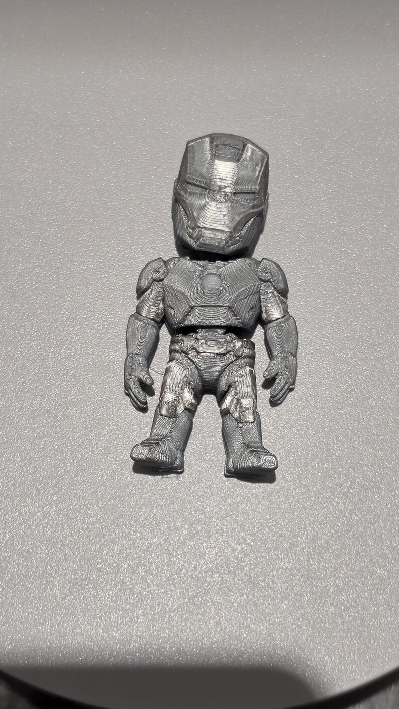 May include: A small, three-dimensional printed figurine of a character wearing a metallic gray armored suit with a full-face helmet, chest plate, and articulated limbs. The surface texture shows visible horizontal layer lines from the printing process.