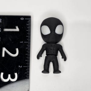K&ouml;nnte beinhalten: Eine schwarze Spider-Man-Figur mit wei&szlig;en Augendetails, etwa 7 cm hoch. Ein schwarzes Lineal mit wei&szlig;en Markierungen befindet sich neben der Figur. Die Figur steht auf einer wei&szlig;en Oberfl&auml;che.