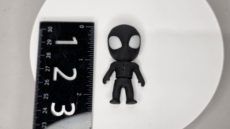 K&ouml;nnte beinhalten: Eine schwarze Spider-Man-Figur mit wei&szlig;en Augendetails, etwa 7 cm hoch. Ein schwarzes Lineal mit wei&szlig;en Markierungen befindet sich neben der Figur. Die Figur steht auf einer wei&szlig;en Oberfl&auml;che.