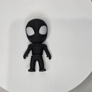 K&ouml;nnte beinhalten: Eine schwarze Superheldenfigur mit wei&szlig;en Augen auf einer wei&szlig;en Oberfl&auml;che. Die Figur hat ein detailliertes Design auf der Brust. Die Figur ist etwa 7 cm gro&szlig;.
