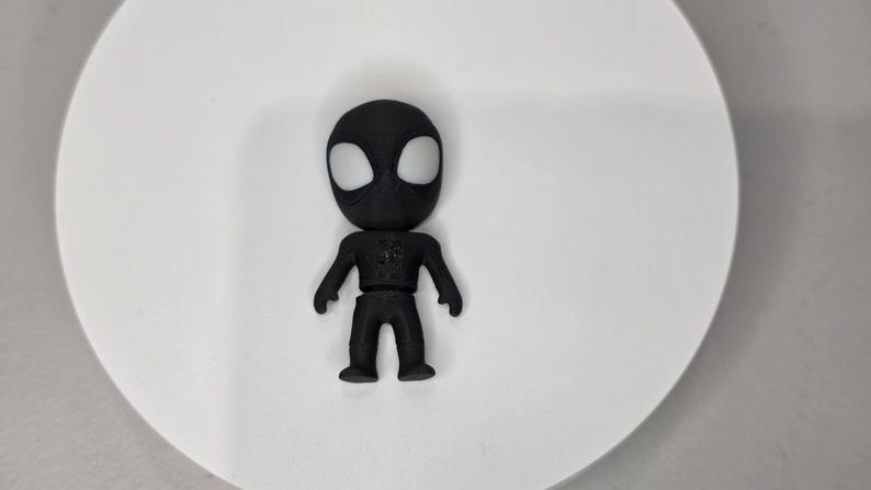 K&ouml;nnte beinhalten: Eine schwarze Superheldenfigur mit wei&szlig;en Augen auf einer wei&szlig;en Oberfl&auml;che. Die Figur hat ein detailliertes Design auf der Brust. Die Figur ist etwa 7 cm gro&szlig;.