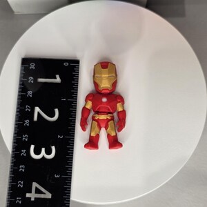 May include: A red and gold Iron Man figurine, approximately 2 inches tall, stands on a white surface. The figurine has a detailed design, with a gold helmet and accents. A black ruler is placed next to the figurine.