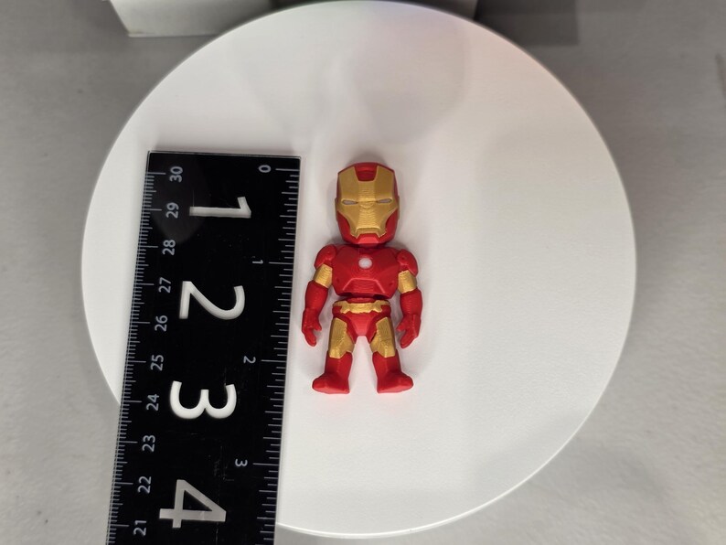 May include: A red and gold Iron Man figurine, approximately 2 inches tall, stands on a white surface. The figurine has a detailed design, with a gold helmet and accents. A black ruler is placed next to the figurine.