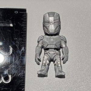 May include: A silver-colored, 3D-printed figurine of a character wearing a full-body armored suit with a helmet. The figurine is positioned next to a black ruler, showing a height of approximately 2.5 inches from head to toe.
