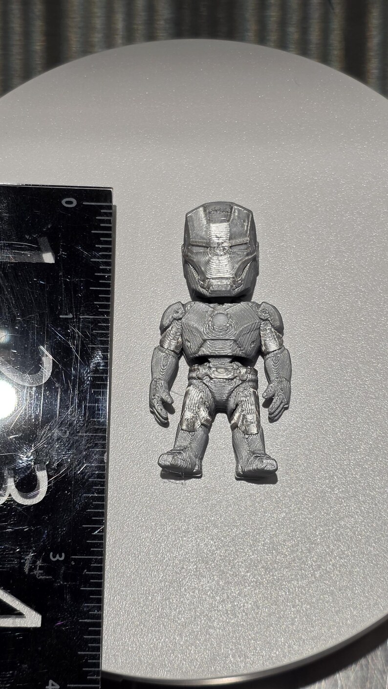 May include: A silver-colored, 3D-printed figurine of a character wearing a full-body armored suit with a helmet. The figurine is positioned next to a black ruler, showing a height of approximately 2.5 inches from head to toe.