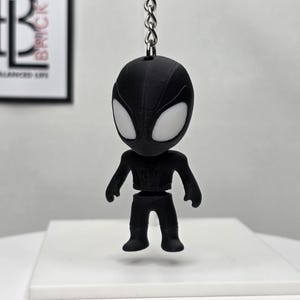 K&ouml;nnte beinhalten: Ein schwarzer Spider-Man-Schl&uuml;sselanh&auml;nger mit wei&szlig;en Augen, der an einer silbernen Kette h&auml;ngt. Die Figur ist stilisiert mit einem gro&szlig;en Kopf und einem kleinen K&ouml;rper. Der Hintergrund enth&auml;lt ein gerahmtes Bild mit dem Text "BALANCED LIFE".