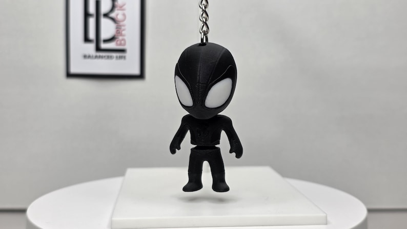 K&ouml;nnte beinhalten: Ein schwarzer Spider-Man-Schl&uuml;sselanh&auml;nger mit wei&szlig;en Augen, der an einer silbernen Kette h&auml;ngt. Die Figur ist stilisiert mit einem gro&szlig;en Kopf und einem kleinen K&ouml;rper. Der Hintergrund enth&auml;lt ein gerahmtes Bild mit dem Text "BALANCED LIFE".