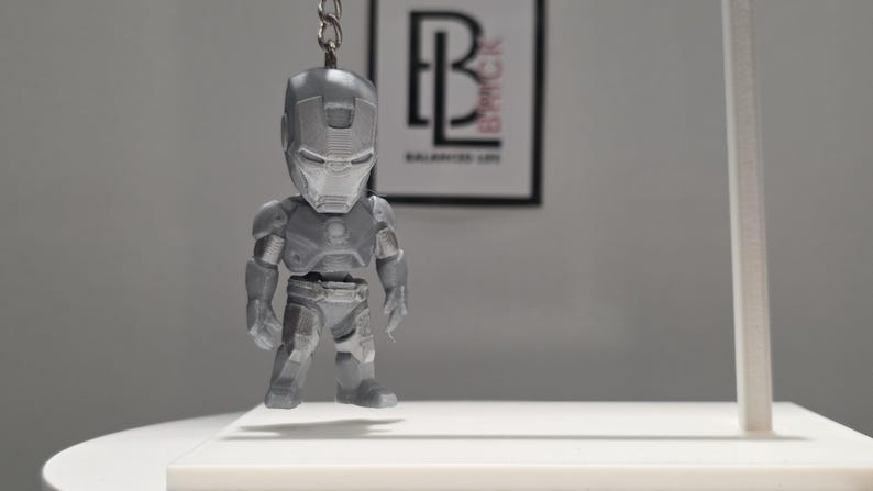 May include: A silver-colored Iron Man character keychain featuring a stylized, chibi-inspired design with a metallic finish. The figure is suspended from a metal chain and ring, showcasing detailed armor plating on the head, torso, arms, and legs.