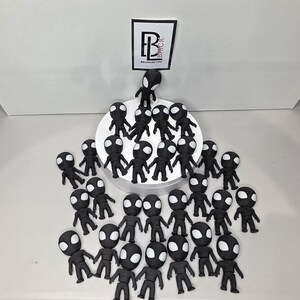 K&ouml;nnte beinhalten: Eine Ansammlung kleiner, schwarzer Figuren mit wei&szlig;en augenf&ouml;rmigen Markierungen. Die Figuren sind auf einer wei&szlig;en Oberfl&auml;che angeordnet, einige stehen auf einer wei&szlig;en, kreisf&ouml;rmigen Plattform. Ein Logo mit dem Text "BALANCED LIFE" ist sichtbar.