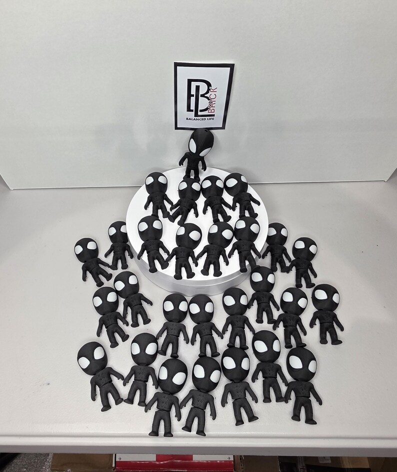 K&ouml;nnte beinhalten: Eine Ansammlung kleiner, schwarzer Figuren mit wei&szlig;en augenf&ouml;rmigen Markierungen. Die Figuren sind auf einer wei&szlig;en Oberfl&auml;che angeordnet, einige stehen auf einer wei&szlig;en, kreisf&ouml;rmigen Plattform. Ein Logo mit dem Text "BALANCED LIFE" ist sichtbar.