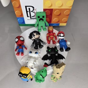 May include: A collection of colorful figurines of popular characters, including Spider-Man, Mario, and a Minion, arranged on a white plate. The figurines are made of plastic and are various sizes. A box with the text "Balanced Brick" is in the background.