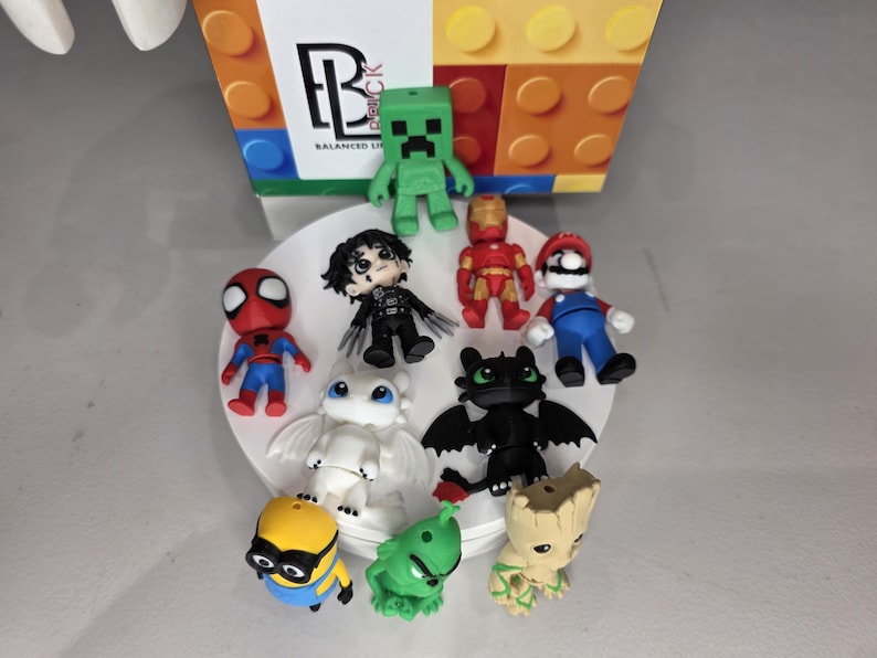 May include: A collection of colorful figurines of popular characters, including Spider-Man, Mario, and a Minion, arranged on a white plate. The figurines are made of plastic and are various sizes. A box with the text "Balanced Brick" is in the background.