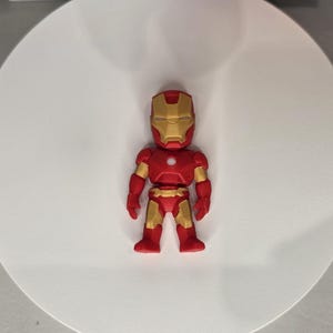 May include: A red and gold Iron Man figurine. The figure has a red body with gold accents on the arms, legs, and helmet. The helmet features a gold faceplate. The figurine is standing on a white surface.