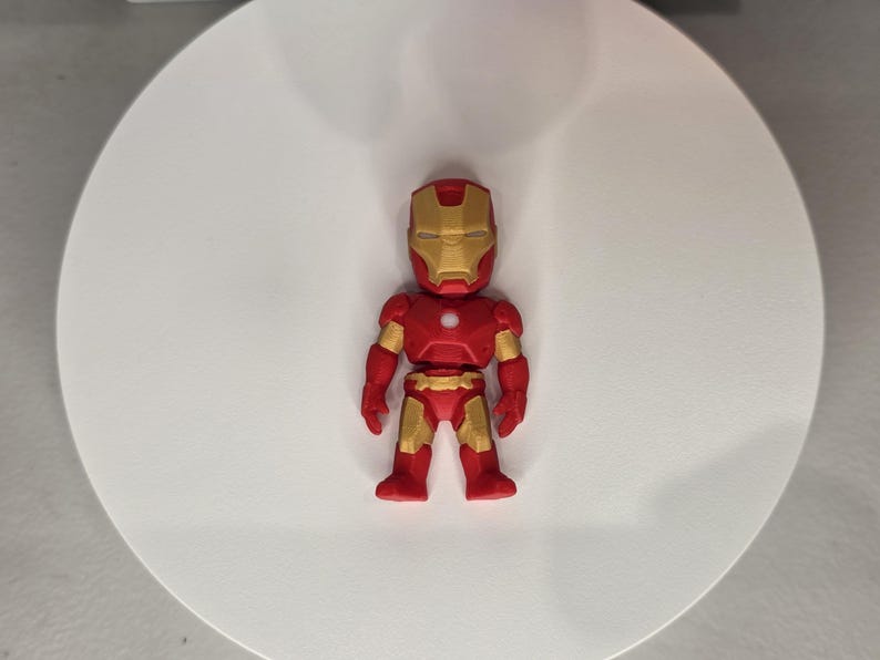 May include: A red and gold Iron Man figurine. The figure has a red body with gold accents on the arms, legs, and helmet. The helmet features a gold faceplate. The figurine is standing on a white surface.