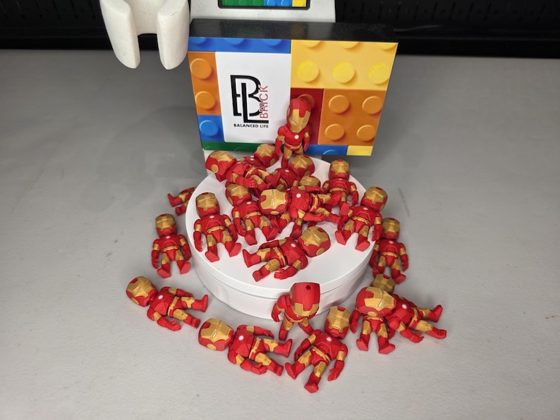 May include: A collection of small, red and gold Iron Man figurines are scattered on a white surface. A colorful box with the text "Balanced Life" and a logo is in the background. The figurines are in various poses.