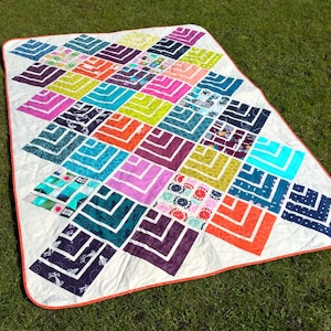 Könnte beinhalten: Ein farbenfroher Quilt mit einem geometrischen Muster aus überlappenden Quadraten und Rechtecken. Der Quilt ist aus verschiedenen Stoffen in Blautönen, Grün, Lila, Rosa, Gelb und Orange gefertigt. Der Quilt ist mit einem weißen Rand und einer korallenfarbenen Einfassung eingefasst.
