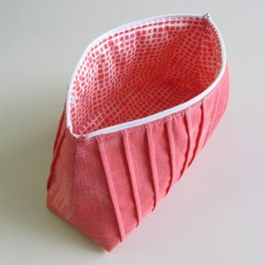 May include: A coral pink fabric zippered pouch with a white and coral pink patterned lining. The pouch is pleated and has a white zipper.